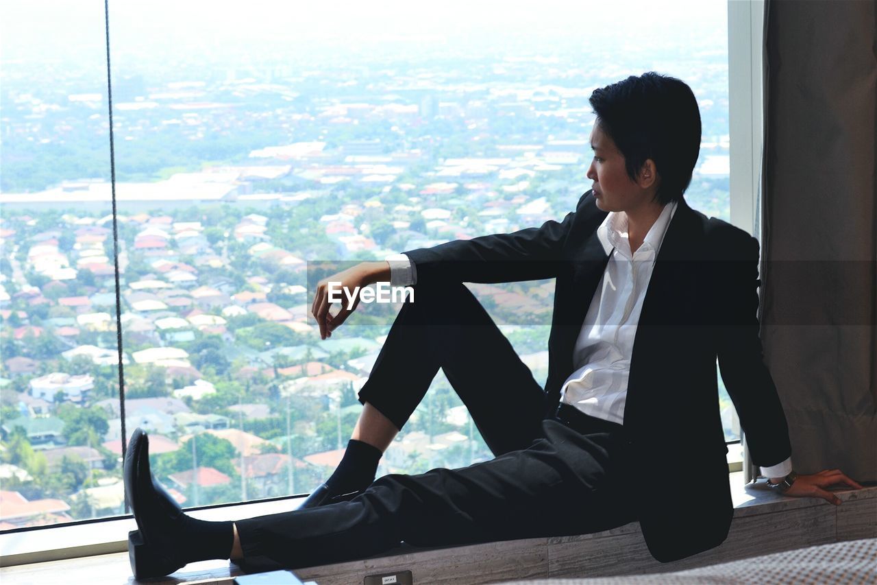 Full length of woman looking at cityscape while sitting by window at home