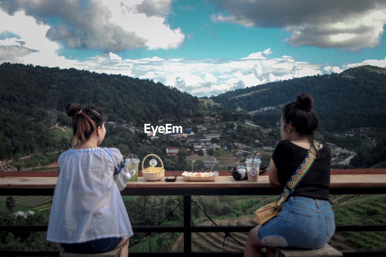 REAR VIEW OF WOMEN SITTING ON TABLE AGAINST MOUNTAIN