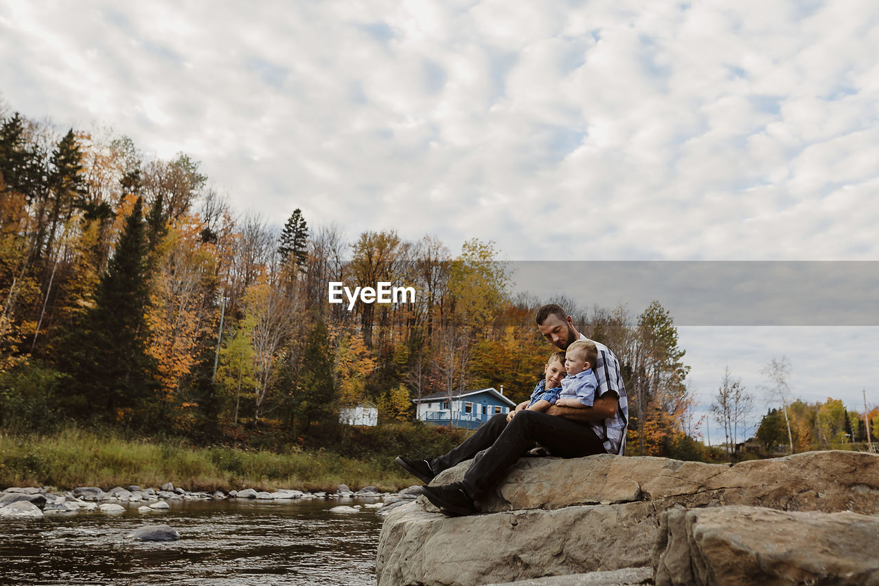 Father with sons sitting on rock by river against cloudy sky in forest