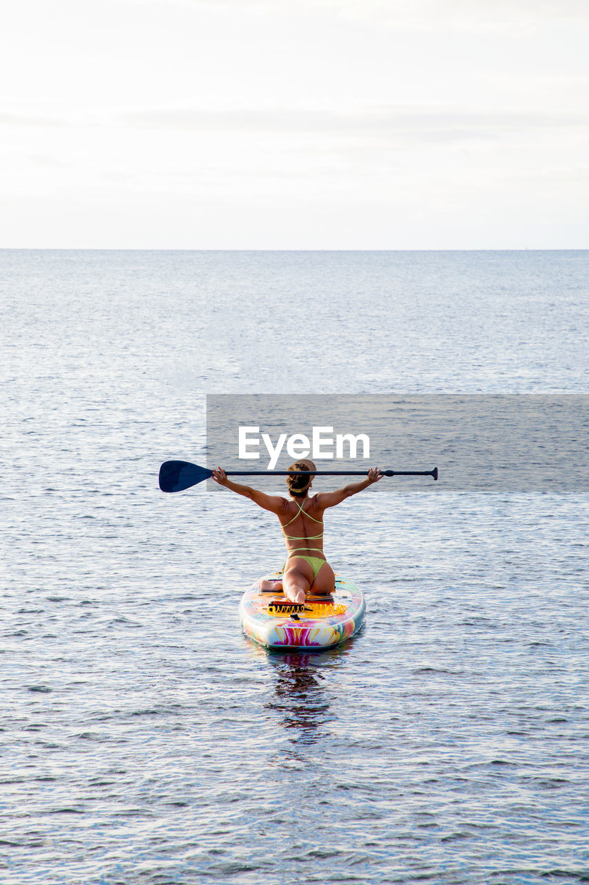 Active female surfer in swimwear holding paddle in raised hand while standing on paddle board floating in sea water near coast