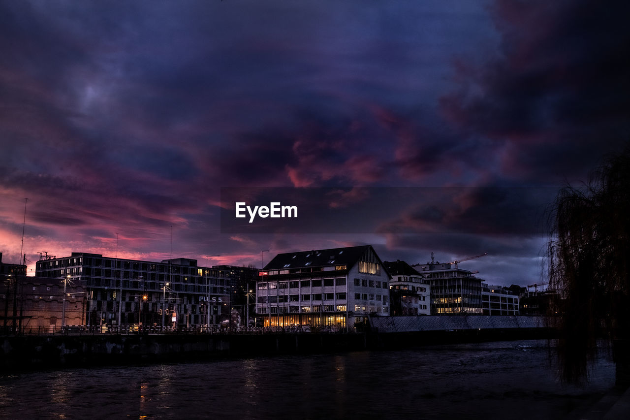 Illuminated buildings by river against sky at sunset