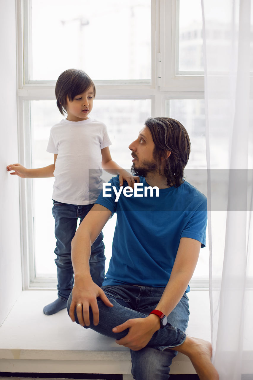 Father in a blue t-shirt and a son in a white t-shirt. sitting on the windowsill of a large window 