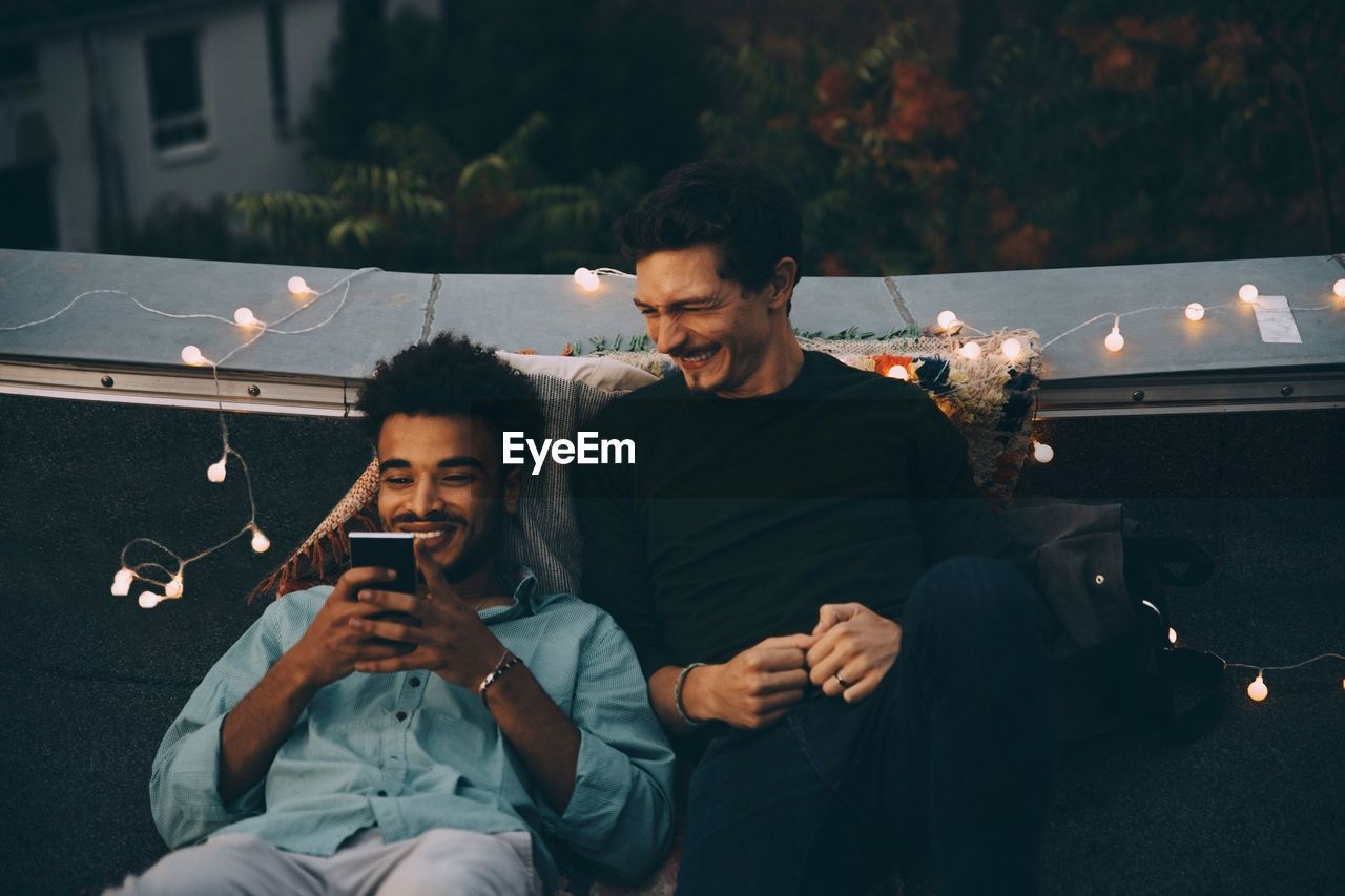 Smiling male friends looking at smart phone sitting on terrace during rooftop party in city