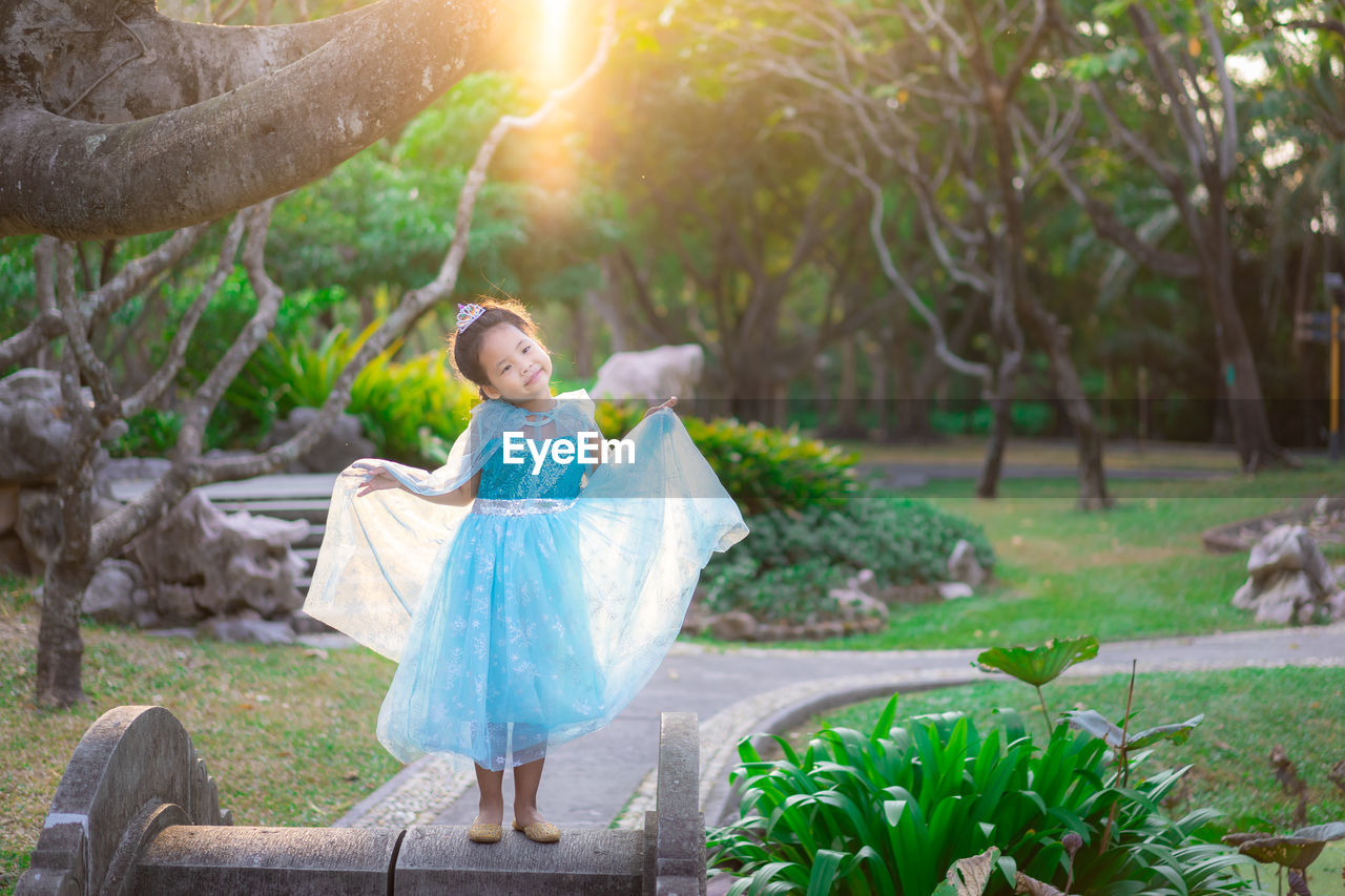 Cute girl wearing blue dress standing against trees in park