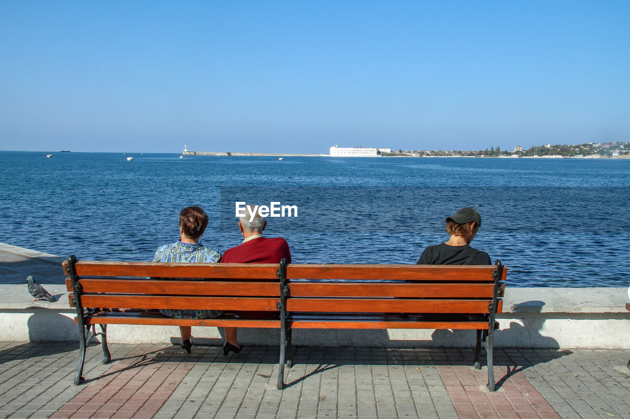 People sitting on bench at beach against clear sky