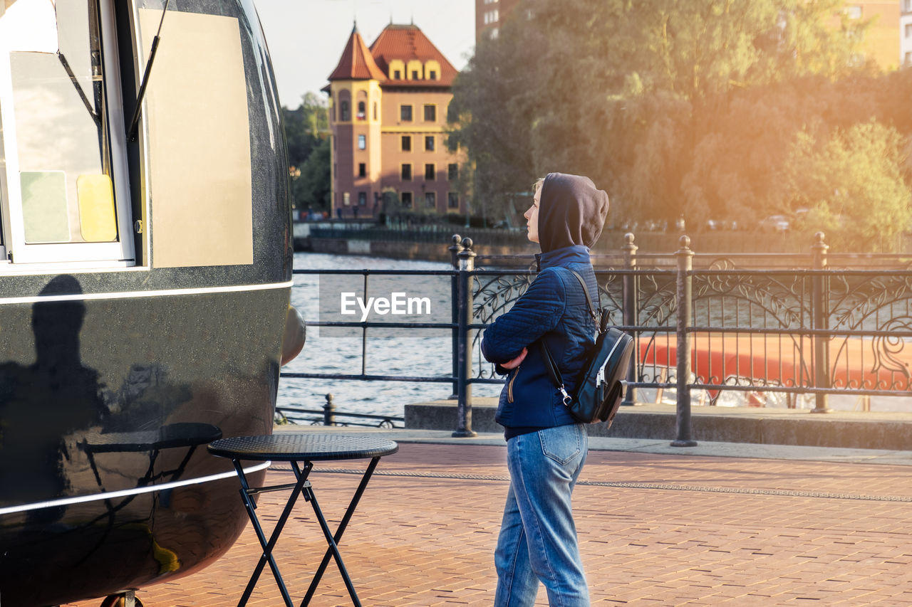 Young woman in hood and with backpack stands near food truck and reads menu. 