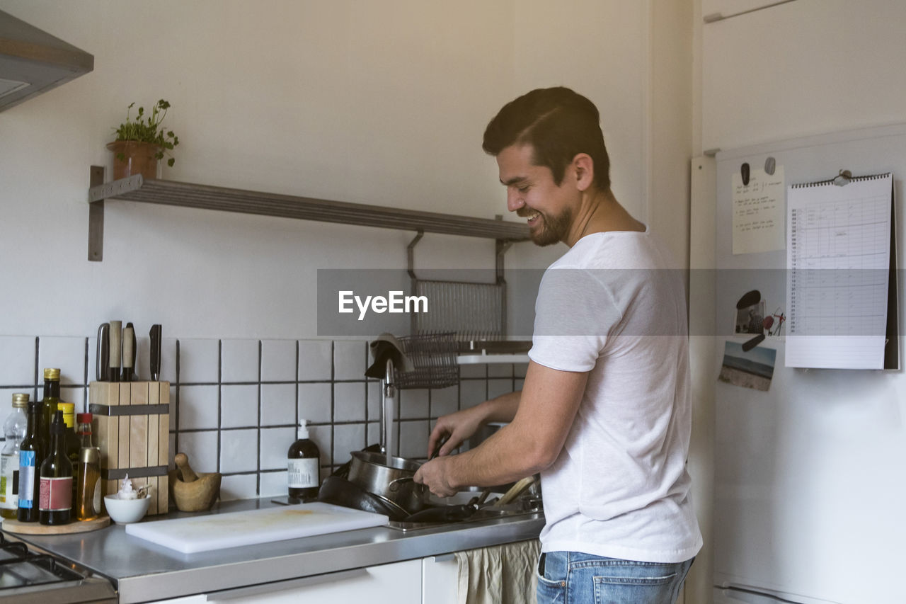 Smiling man washing utensils while standing by sink at kitchen