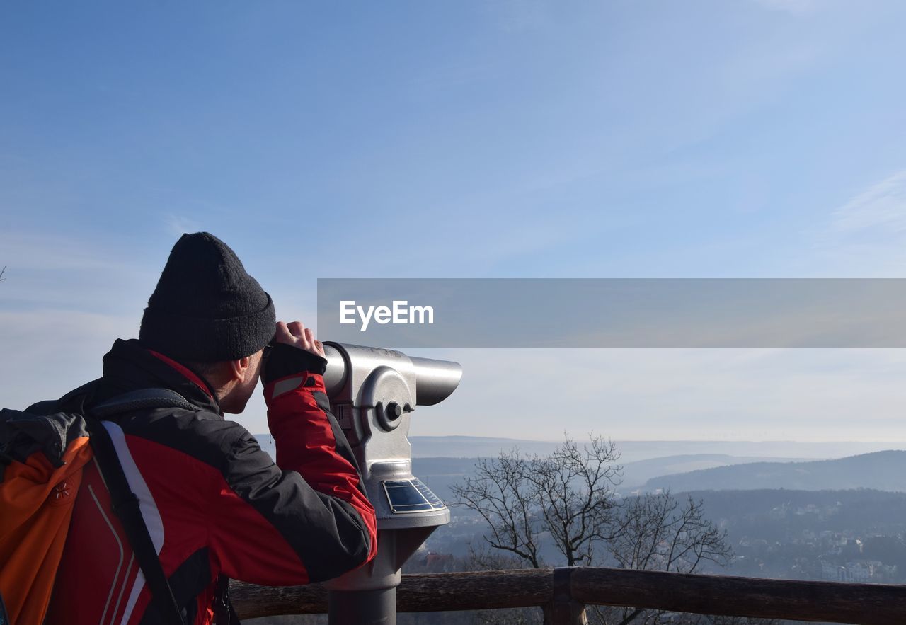 Man looking through coin-operated binoculars against sky during winter