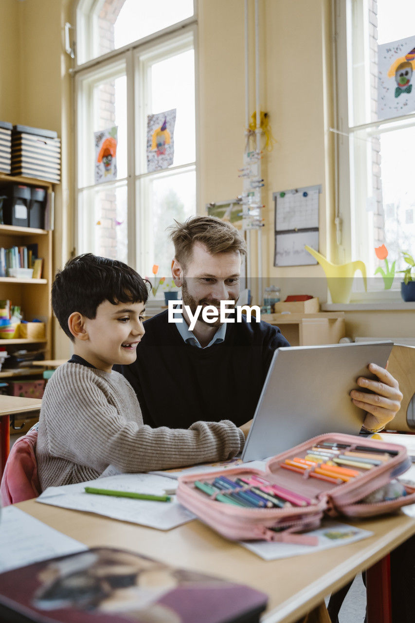 Happy male teacher assisting schoolboy using tablet pc at desk in school