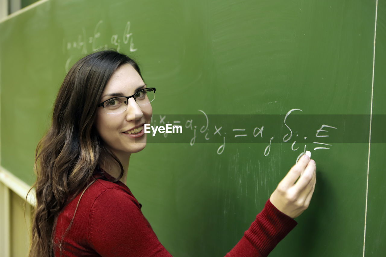 blackboard, board, education, school, classroom, learning, student, school building, women, building, teacher, smiling, intelligence, indoors, eyeglasses, green, portrait, university, glasses, teaching, one person, young adult, adult, architecture, long hair, studying, happiness, university student, emotion, hairstyle, mathematics, standing, brown hair, child, holding, solution, formula, science, teenager, female, waist up, looking at camera, person, headshot, cheerful