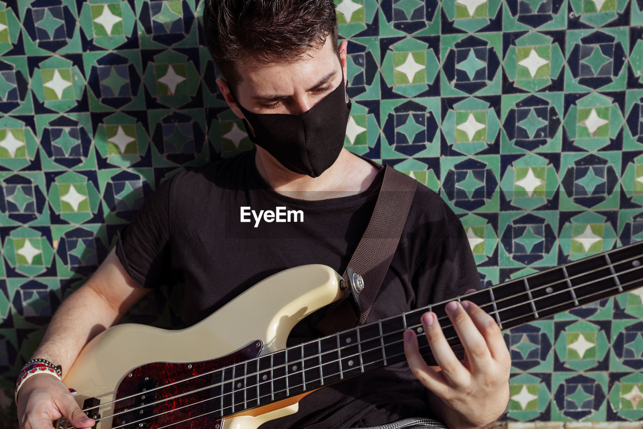 Full body of young man in black protective mask for coronavirus prevention sitting on steps near wall and playing bass guitar on street