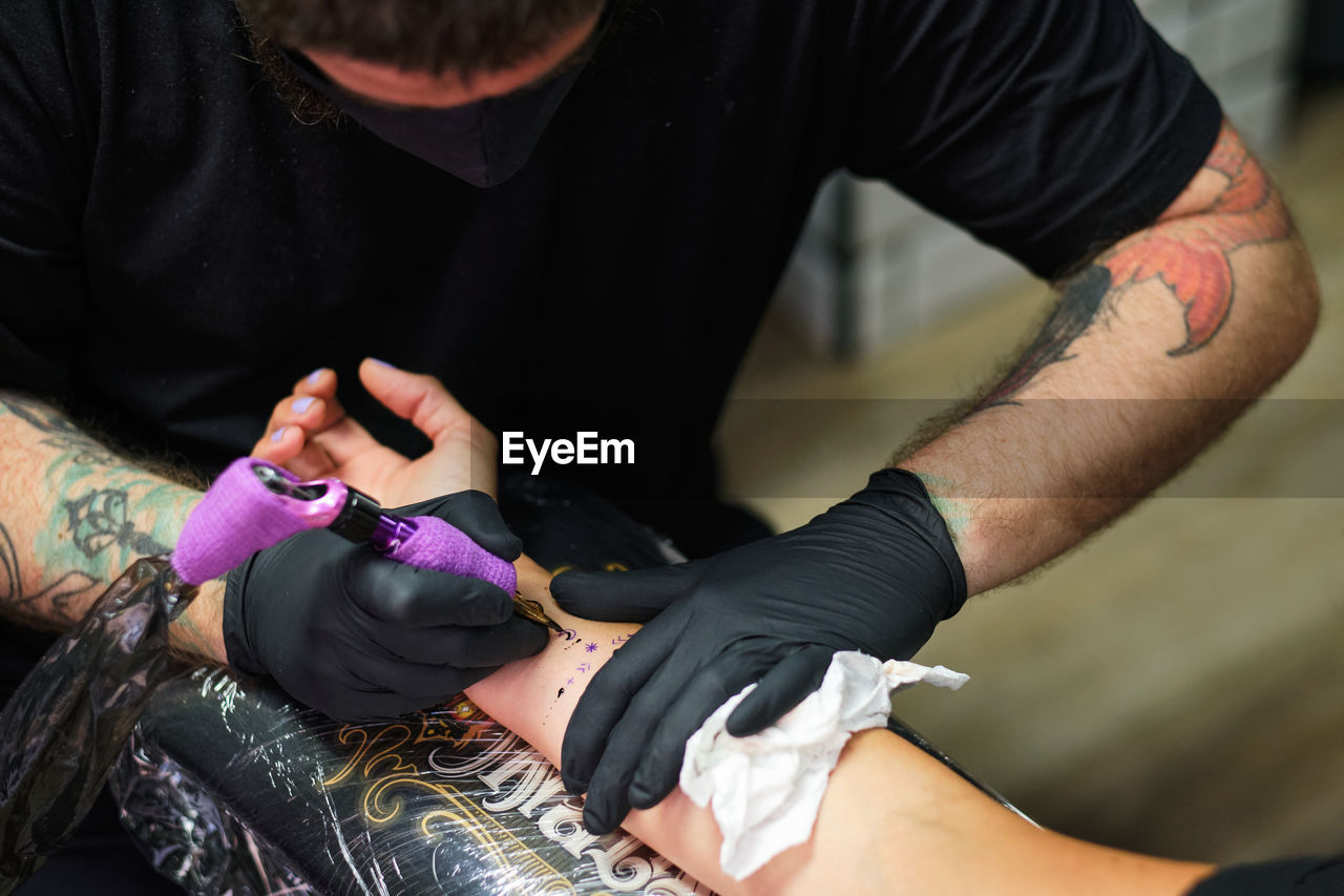 Unrecognizable concentrate bearded male master in black face mask and gloves creating tattoo on arm of crop faceless client in salon