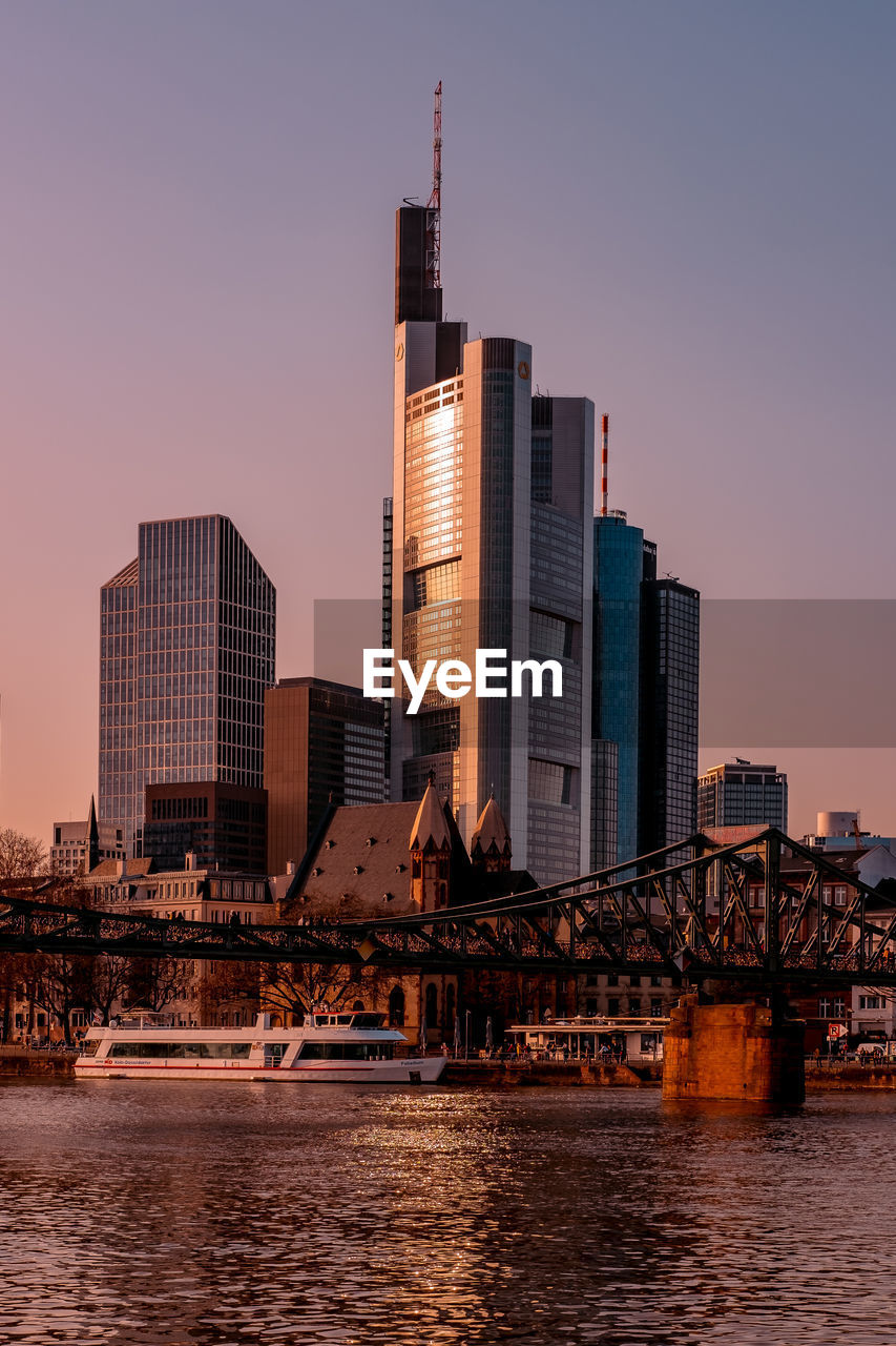 Modern buildings by river against sky during sunset