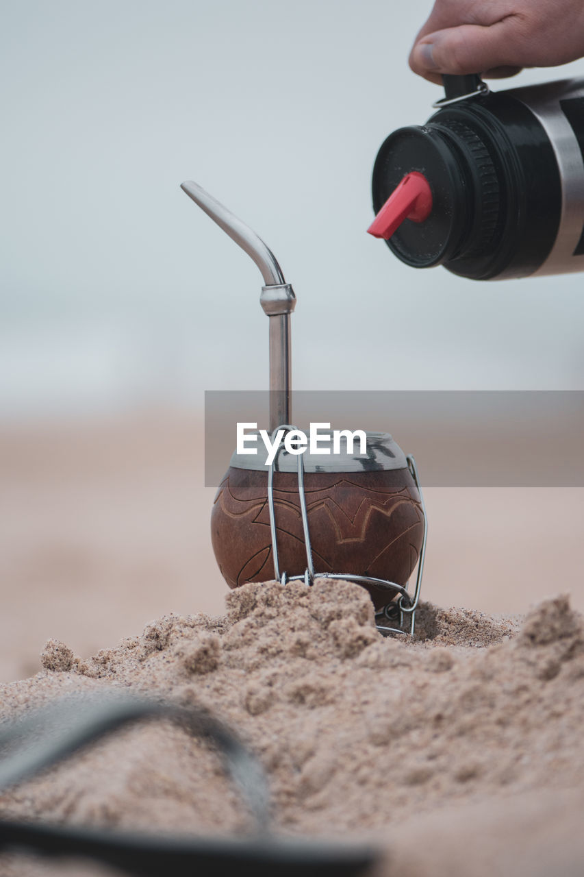 Cropped hand pouring drink in container on sand at beach