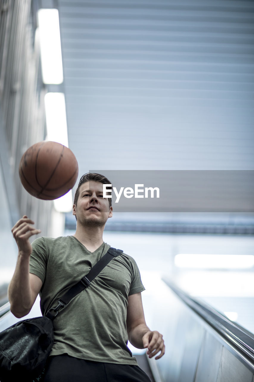 Low angle view of young man playing with basketball while moving down on escalator