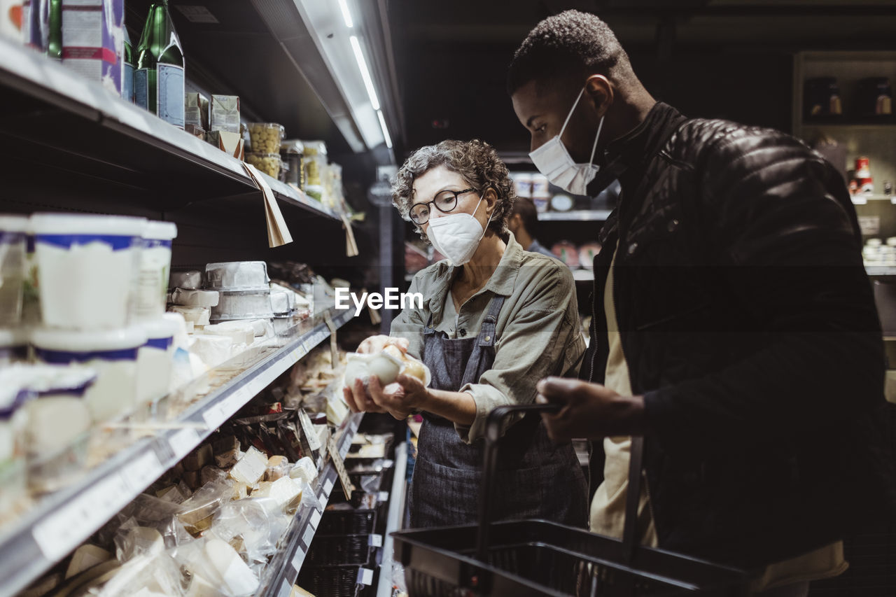 Female owner holding product while male customer standing by in deli store during covid-19