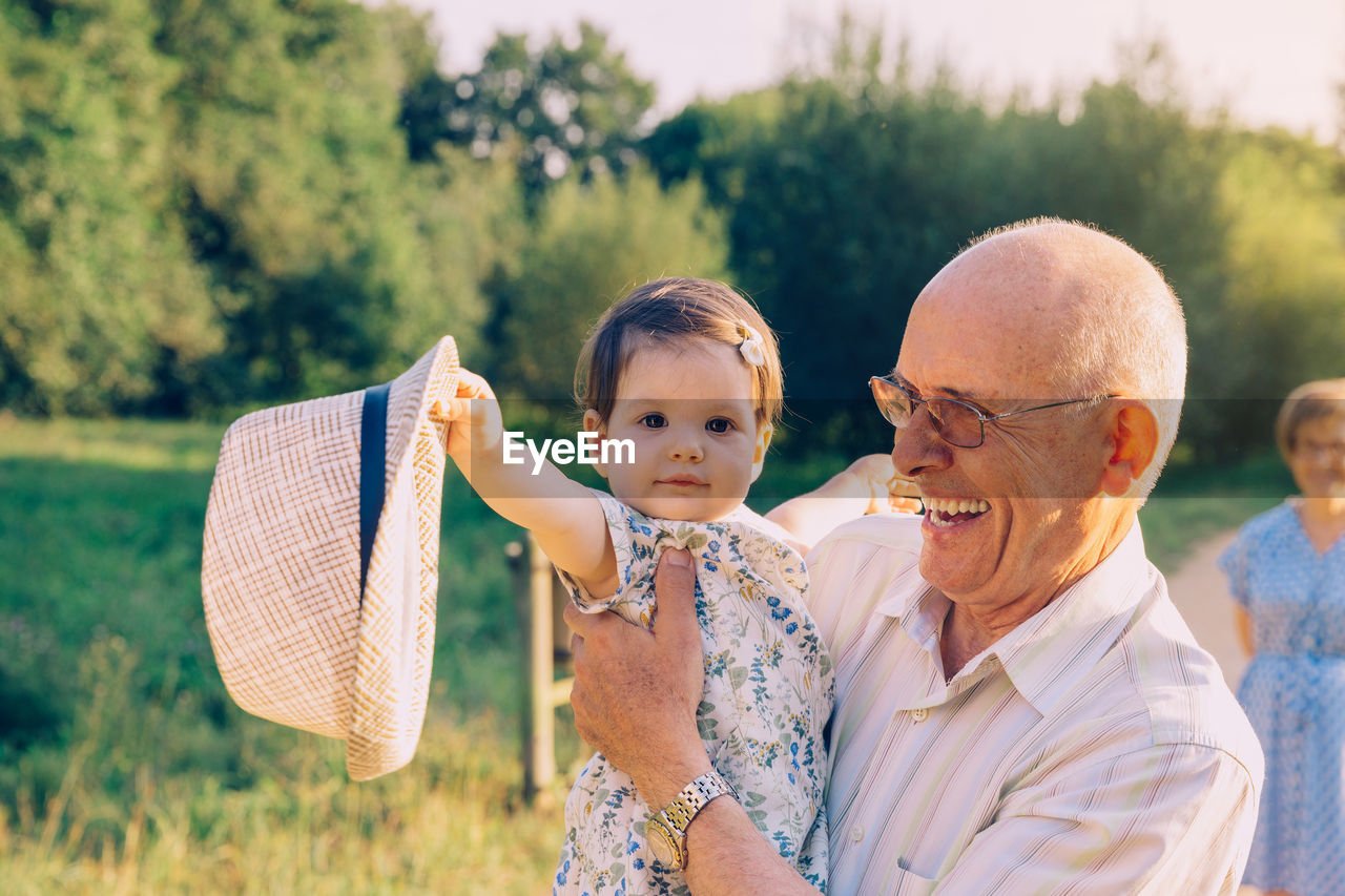 Portrait of granddaughter holding hat with grandfather at park