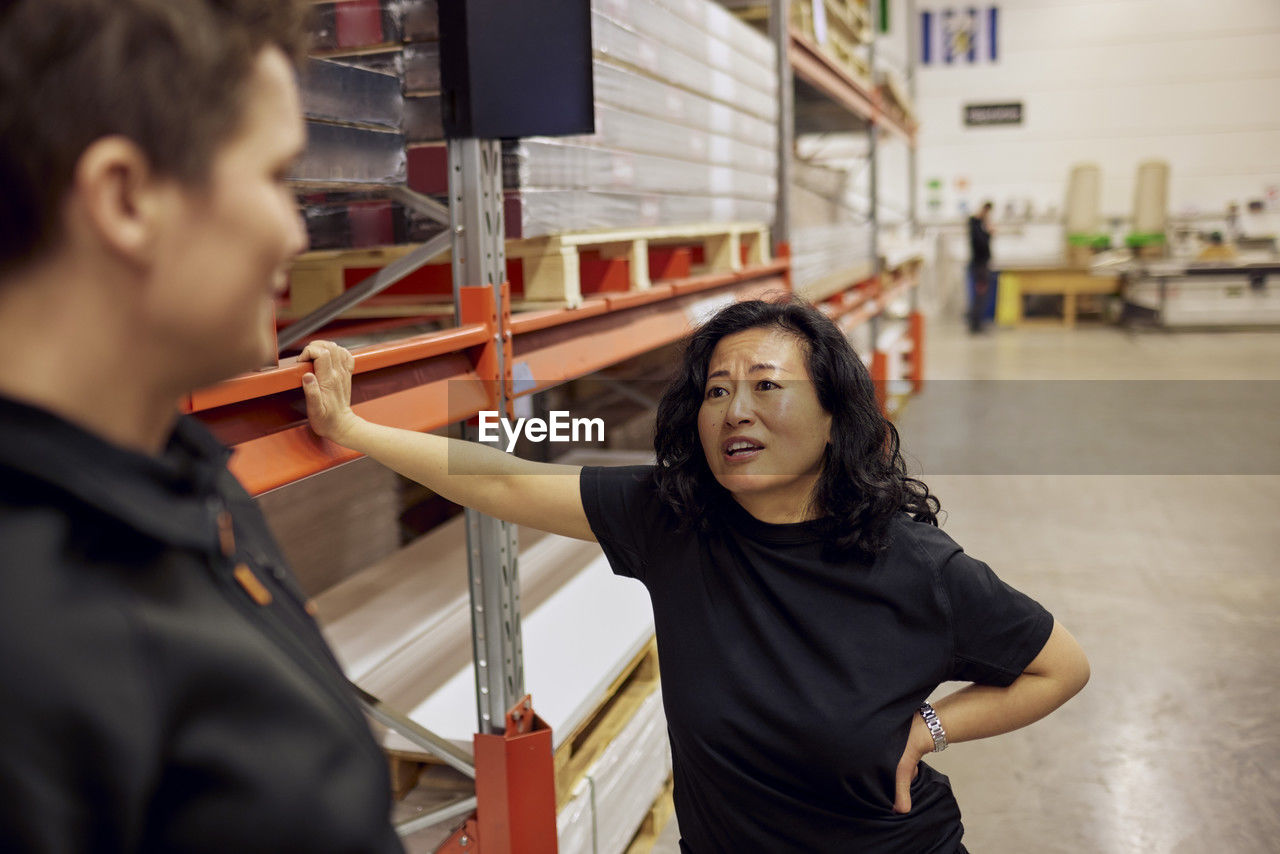Mature female blue-collar worker discussing with colleague while standing by rack in distribution warehouse