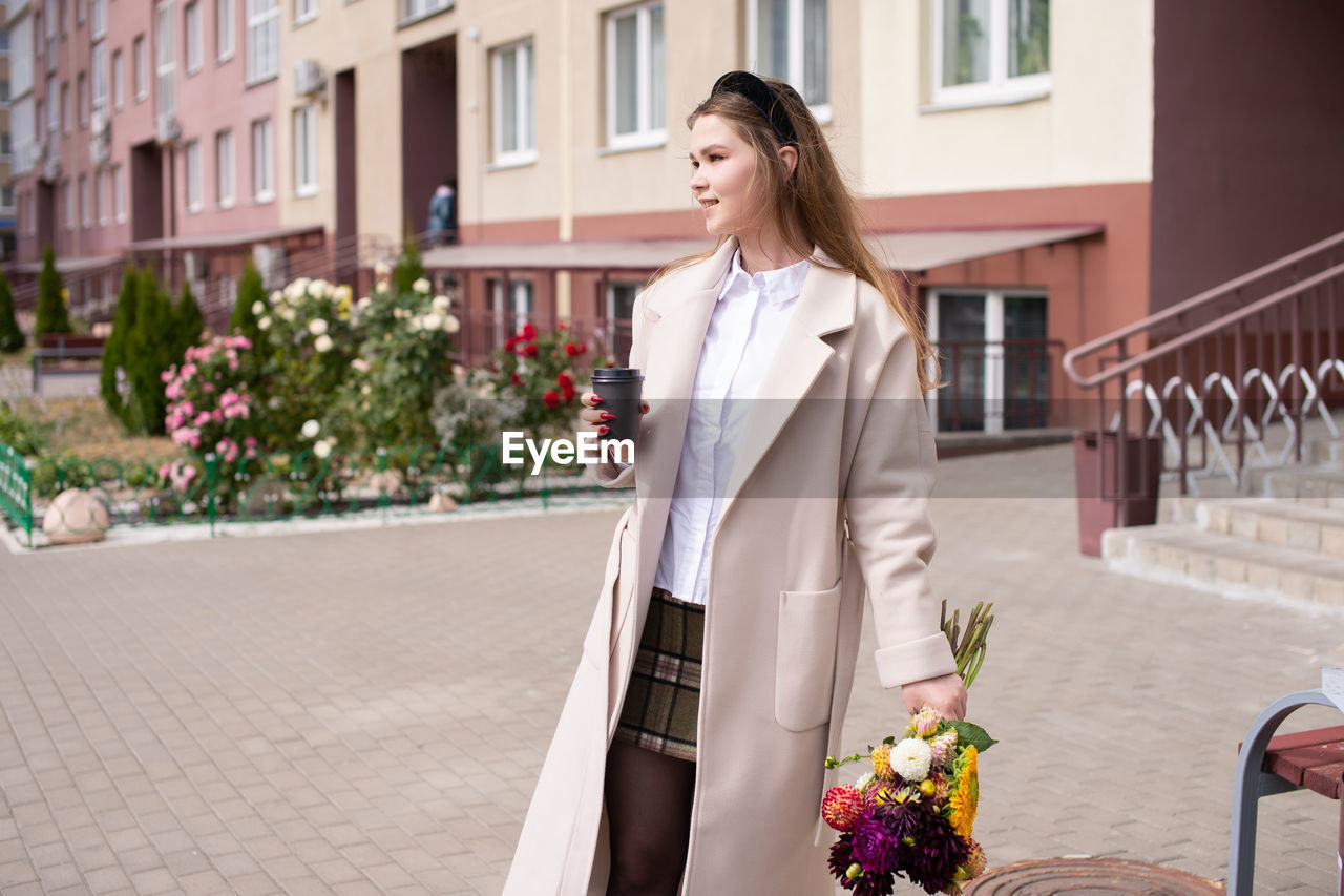 A cute girl in a coat holds a glass of coffee and a bouquet of flowers on the street.