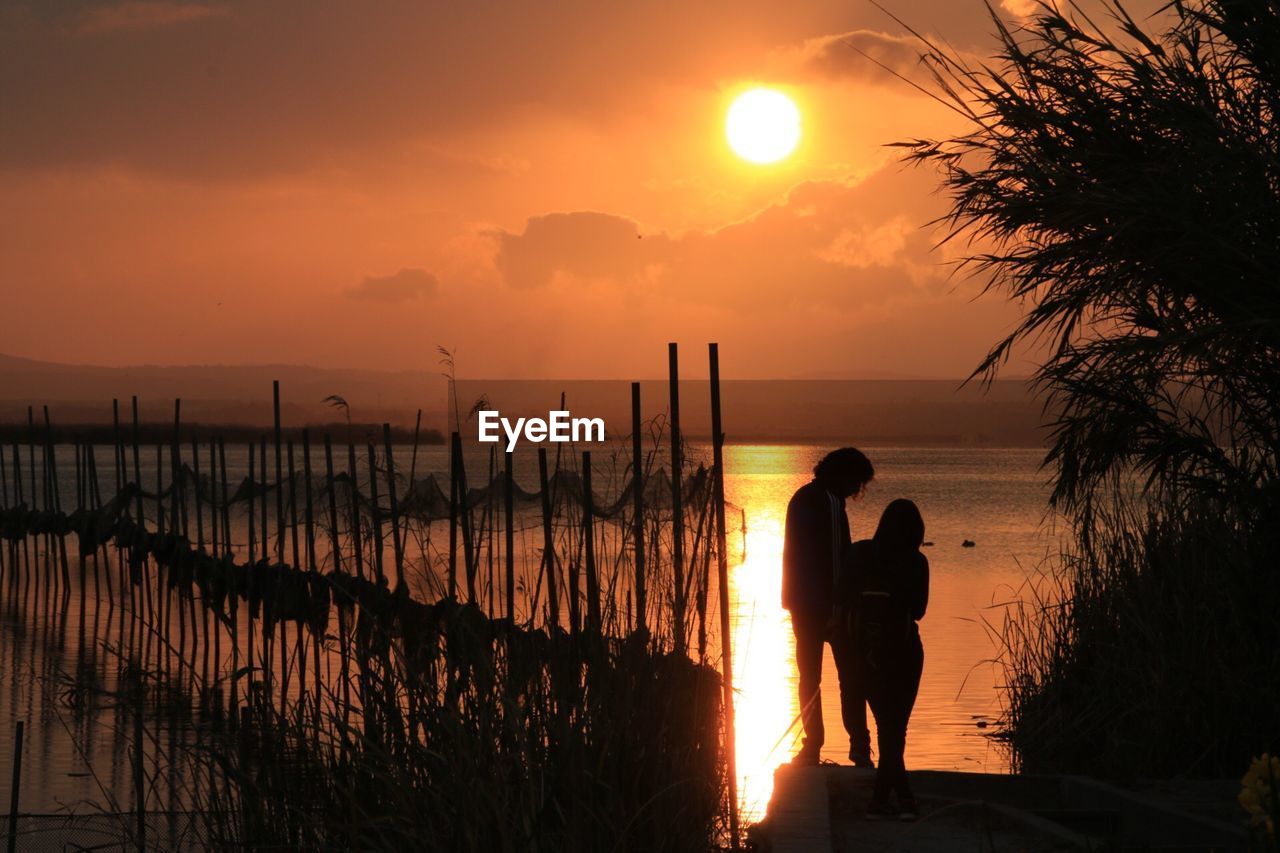 Silhouette man and woman standing by river against sky during sunset