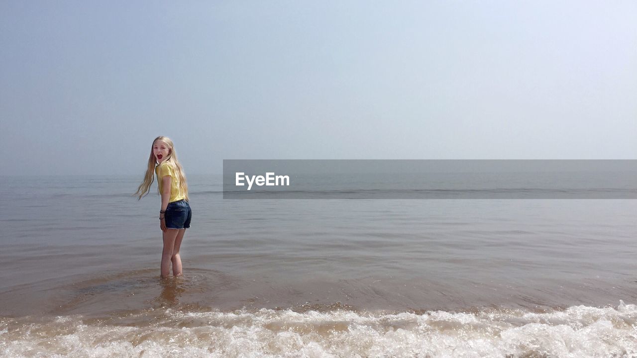 Surprised girl standing at beach against sky