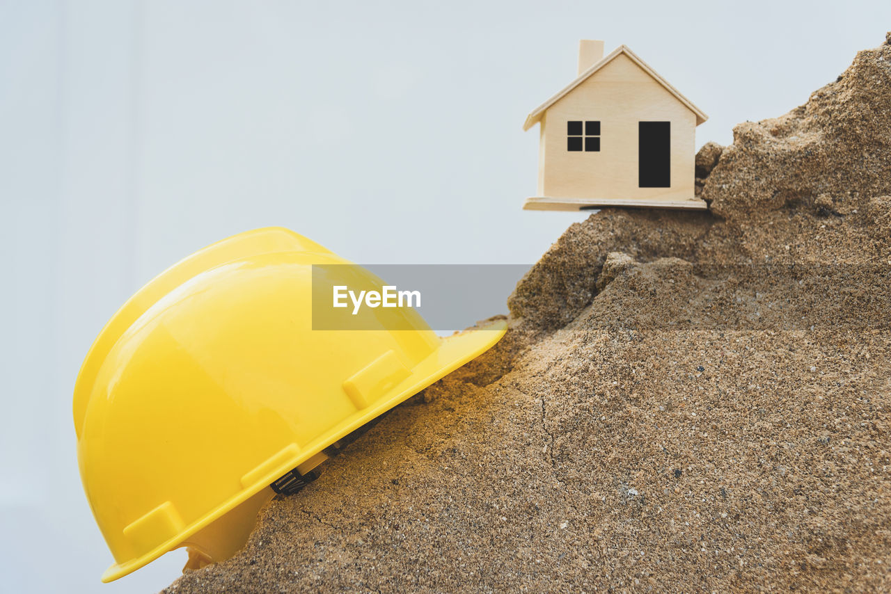 Close-up of yellow hardhat with model home on rock formation against sky