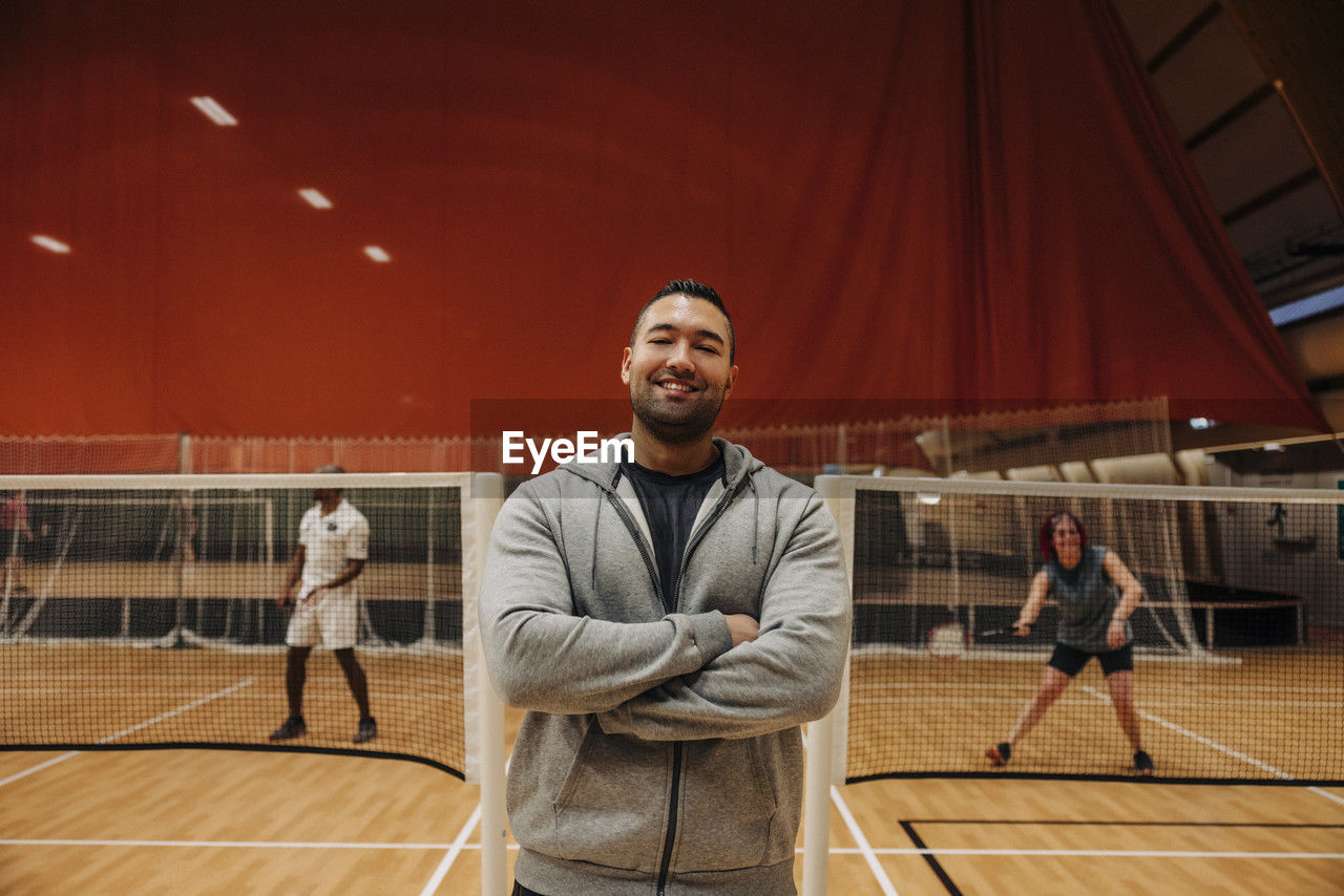 Smiling male coach standing with arms crossed at badminton court