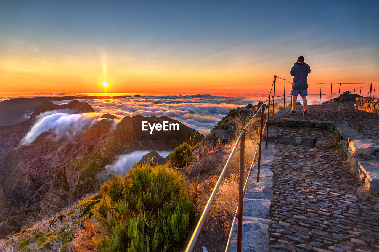 REAR VIEW OF MAN STANDING ON SHORE AGAINST SKY DURING SUNSET