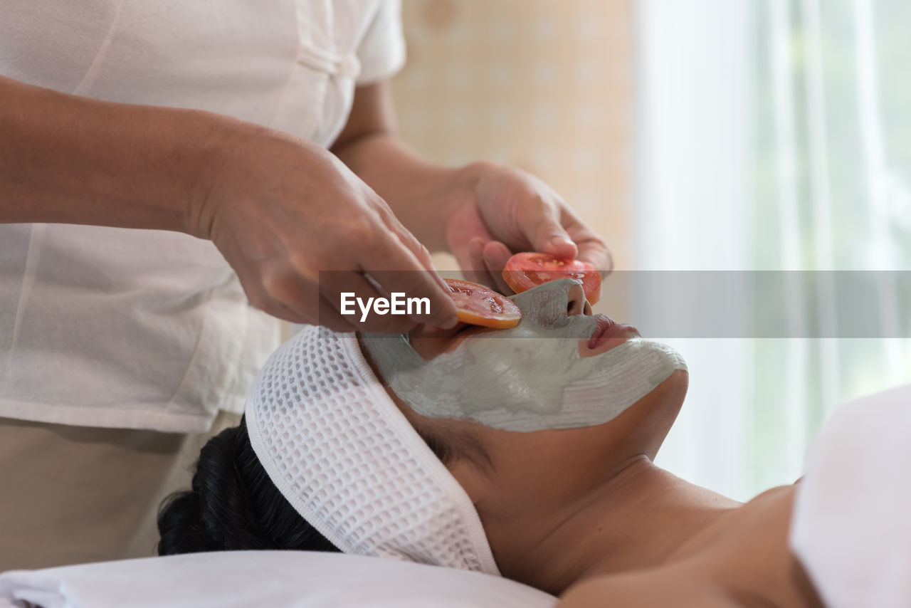 Midsection of beautician adjusting tomato slices on woman eyes during facial mask at spa