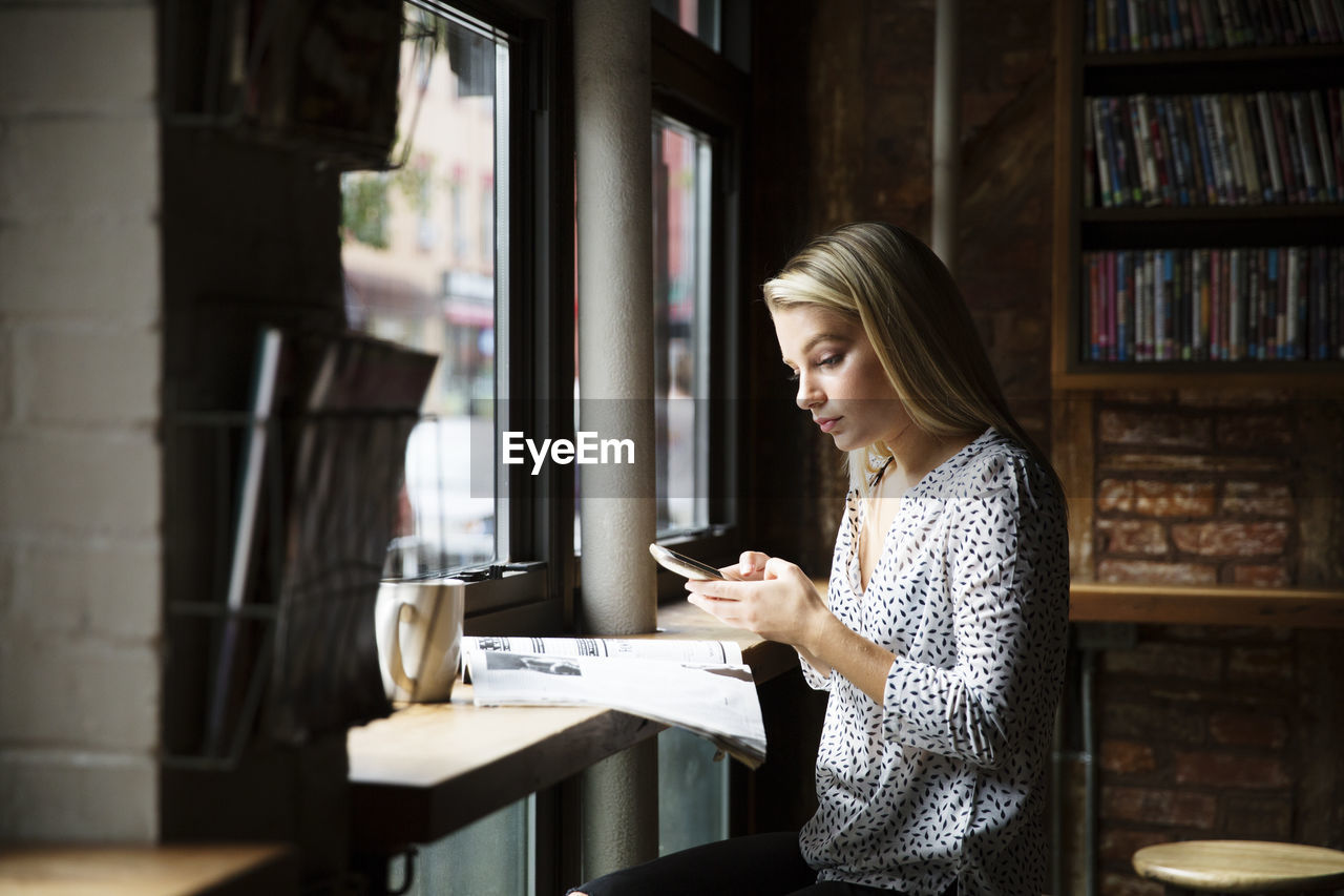 Young woman using phone while reading newspaper at table in cafe