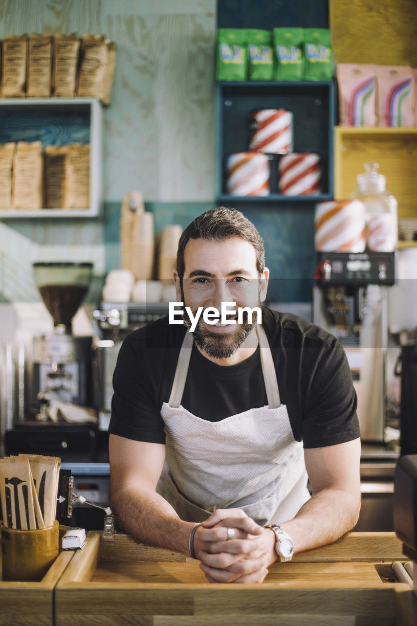 Portrait of smiling male retail clerk with hands clasped leaning on checkout counter at grocery store