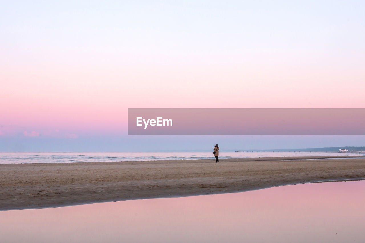 A photographer stands on the wide deserted sea beach surrounded by pink evening light