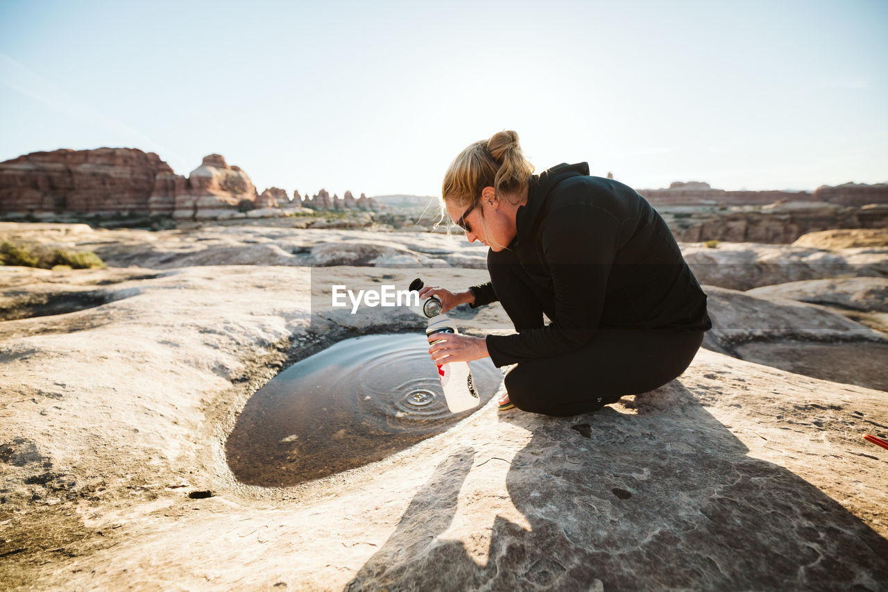 Woman in all black scoops drinking water from a puddle in the desert