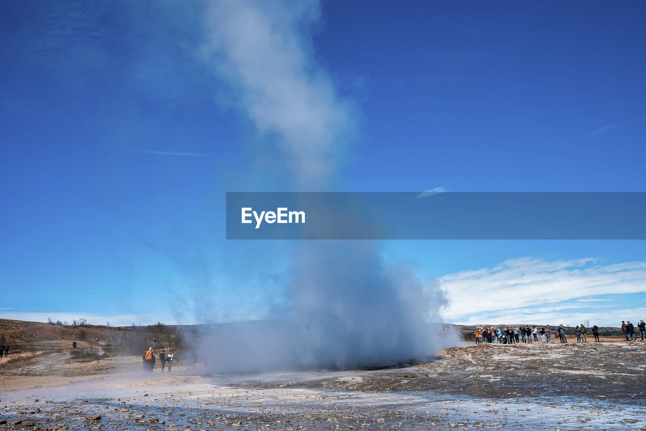 Explorers looking at strokkur geyser eruption in valley against blue sky