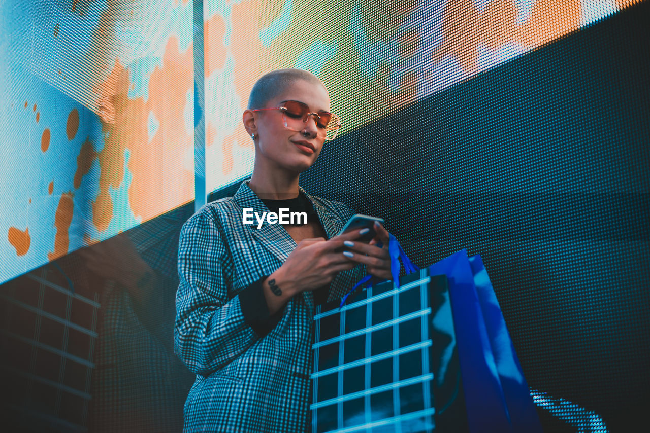 Low angle view of lesbian woman using smart phone holding shopping bags standing against abstract backgrounds