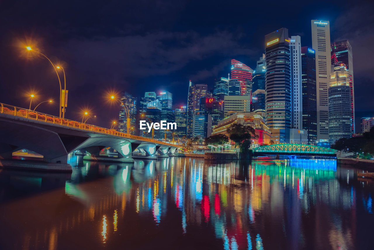 Illuminated bridge over river by buildings against sky at night