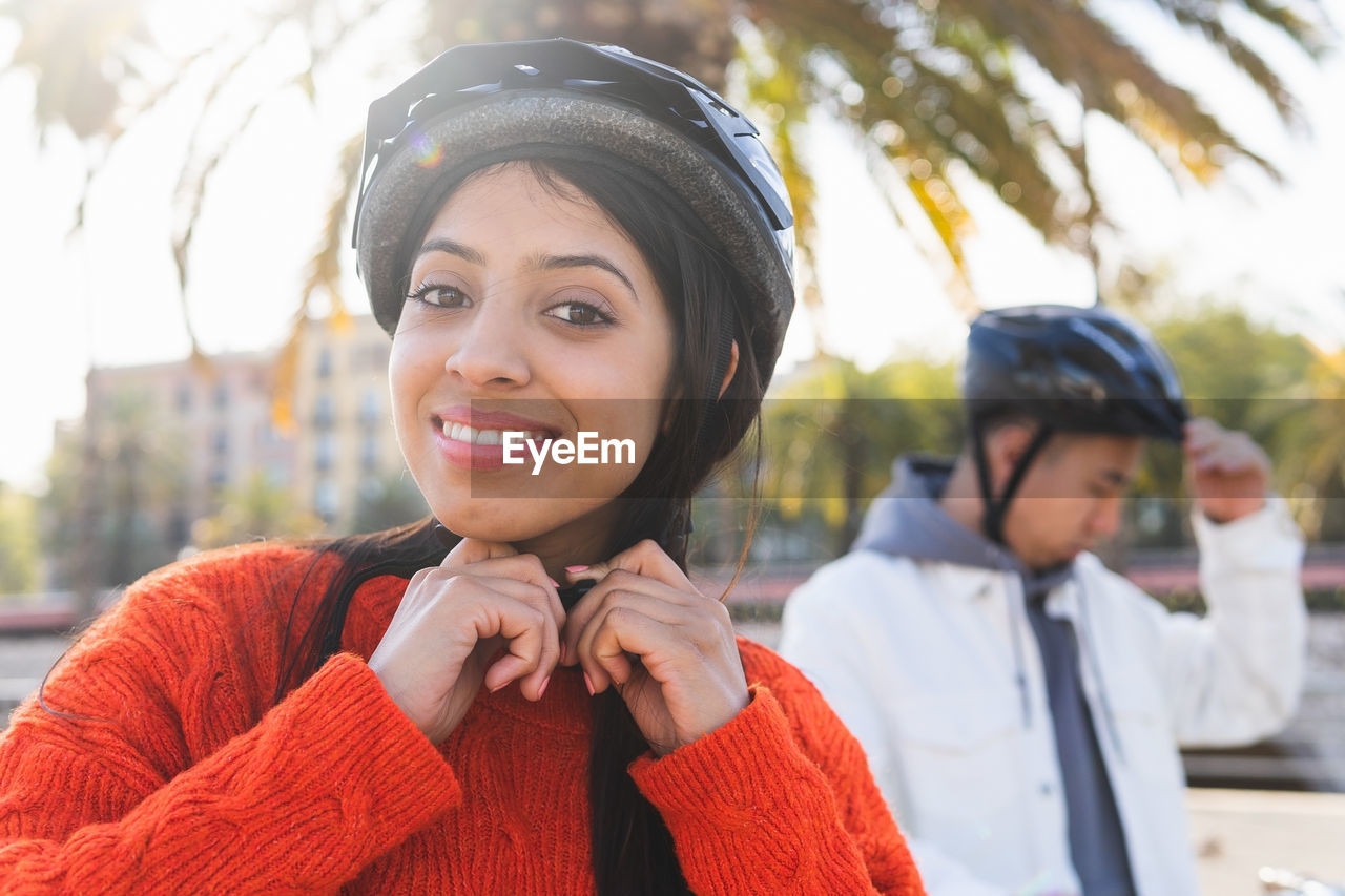 Positive indian girlfriend fastening protective helmet near asian boyfriend while standing together on street with green trees