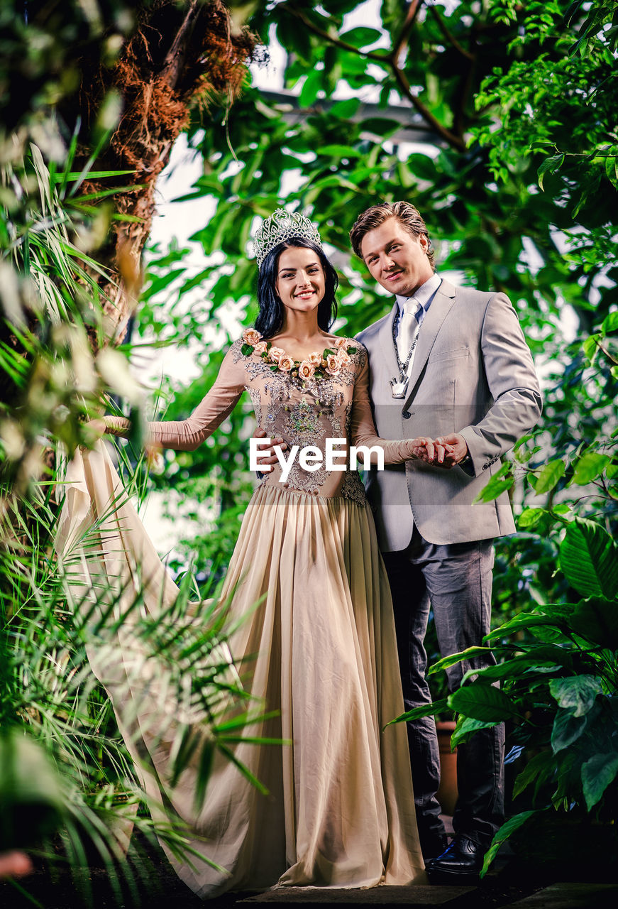 Portrait of smiling bride and groom holding hands while standing amidst plants