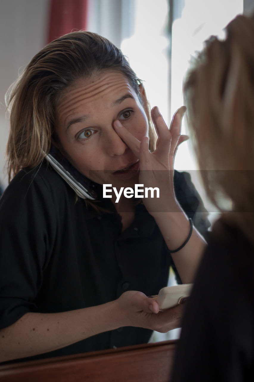 Reflection of woman applying powder compact while talking on phone on mirror