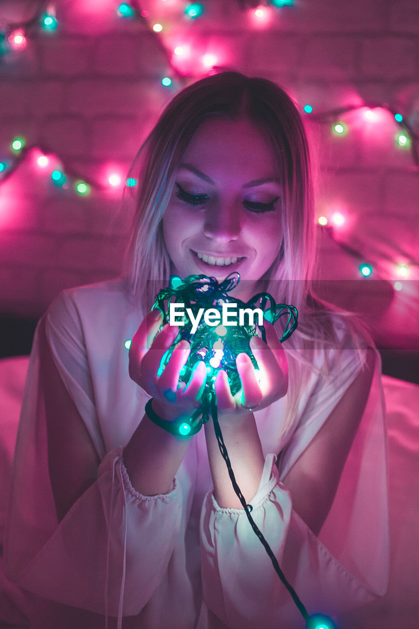 Smiling young woman holding illuminated string lights during christmas