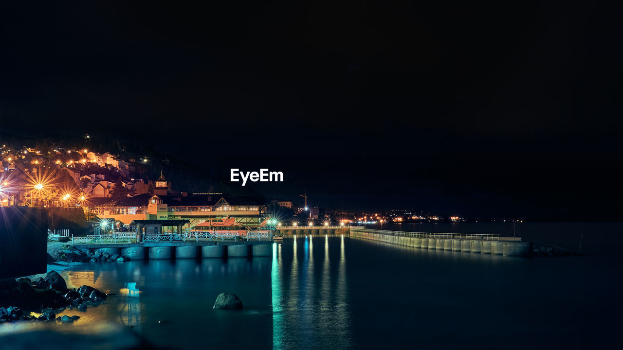 Illuminated city by sea against sky at night