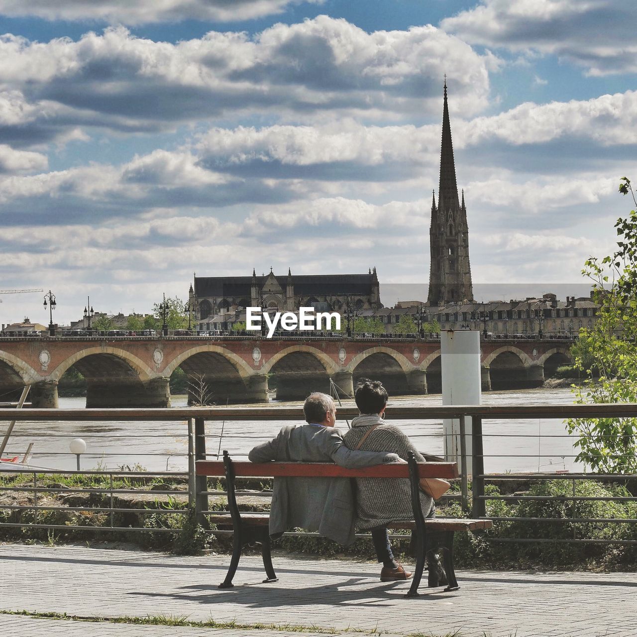 Rear view of man and woman sitting on bench against river in city