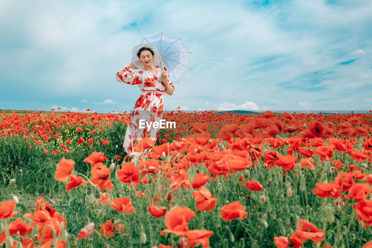 Happy woman in summer dress with a umbrella dances in the field with blooming poppies against a sky