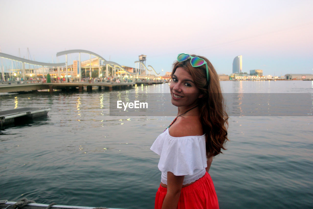 Portrait of beautiful woman standing by river against sky during sunset