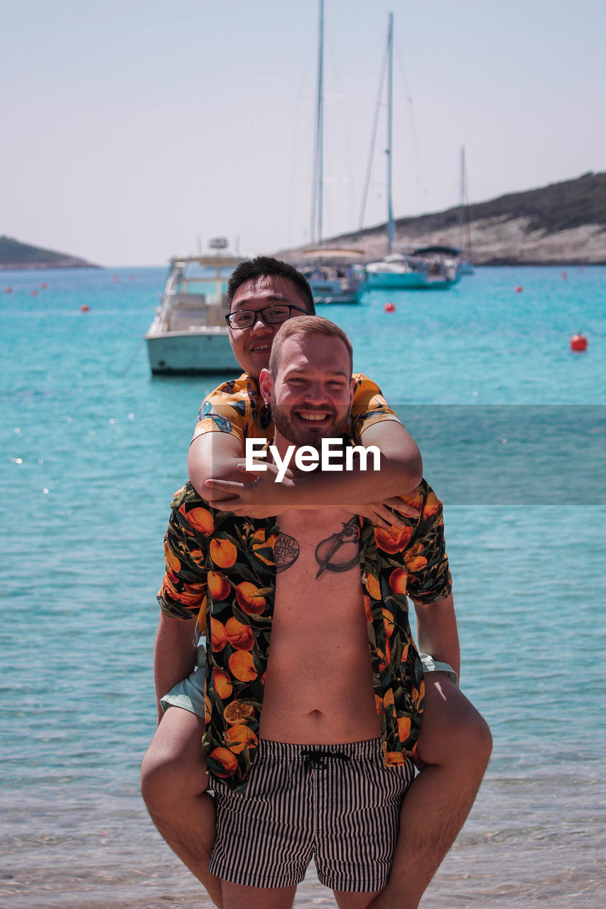 Portrait of happy homosexual couple at beach against clear sky