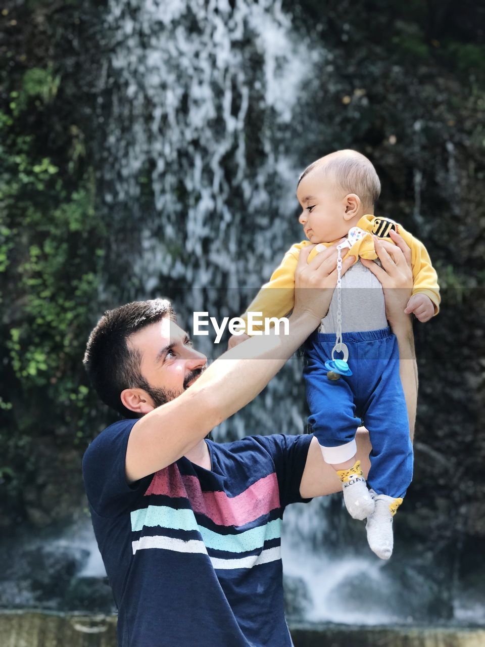 Father picking up son while standing against waterfall