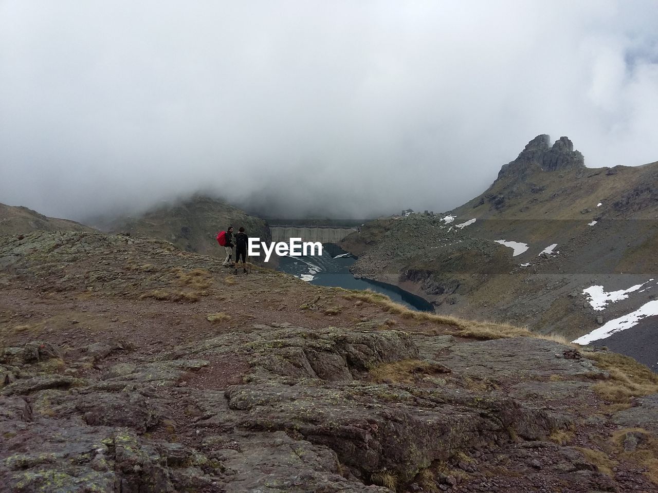 Hikers standing on rocky mountains by dam during foggy weather