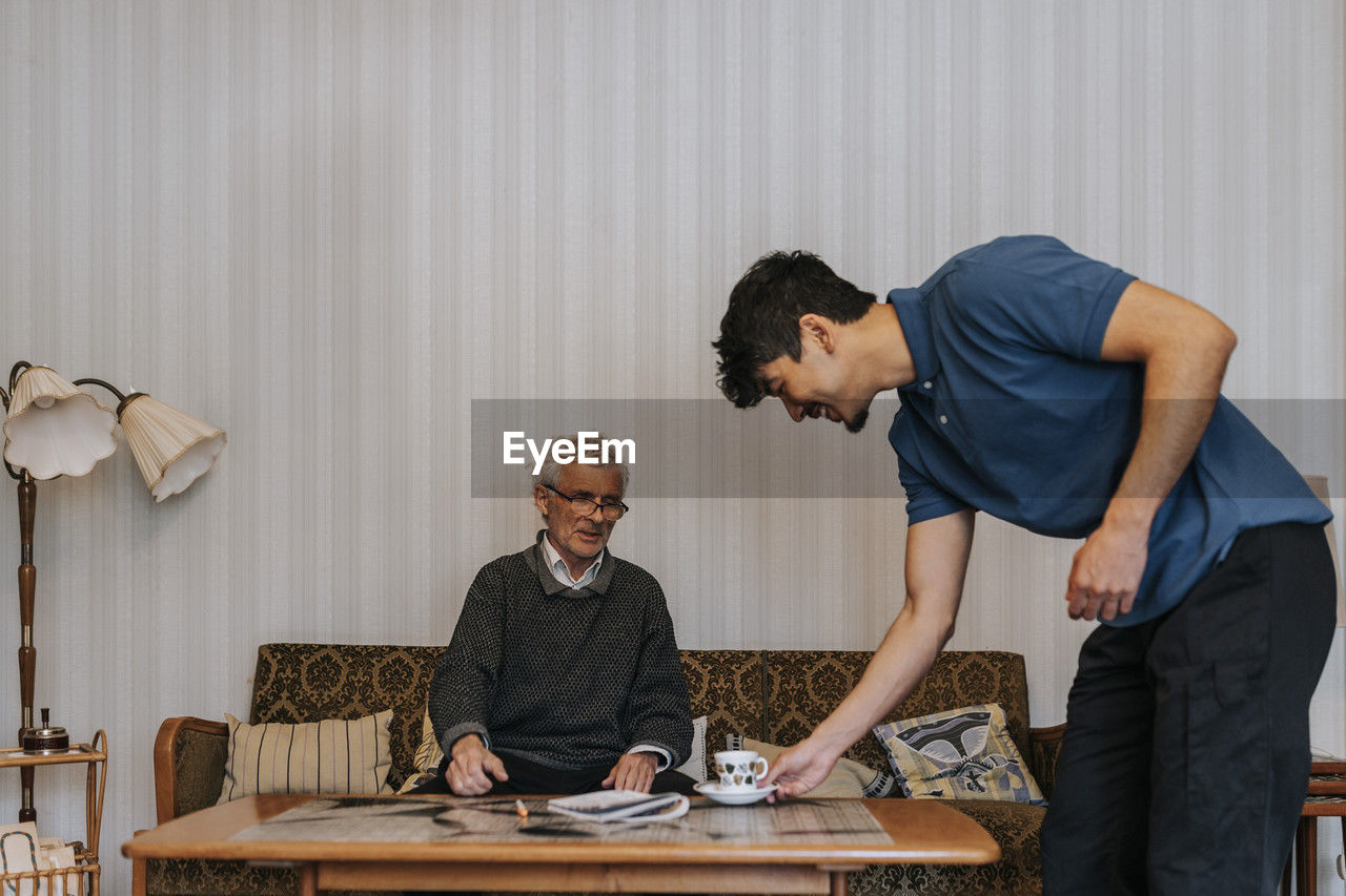 Smiling male care assistant serving tea to senior man sitting on sofa at home