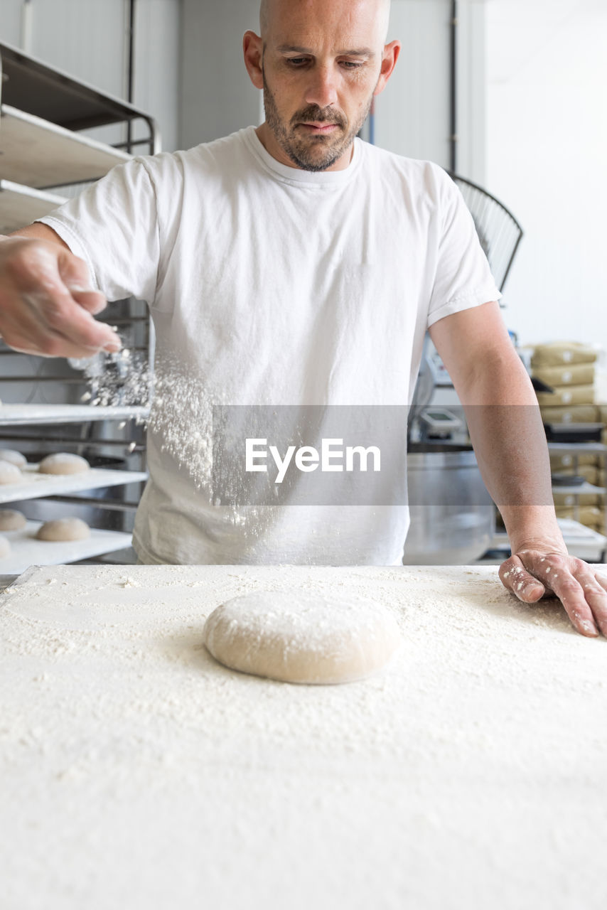 Male worker sprinkling white flour on unbaked dough ball on table while working in light professional bakery