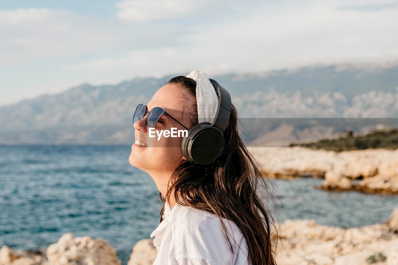 Side view of young woman in white shirt listening to music on headphones. summer, beach, lifestyle.